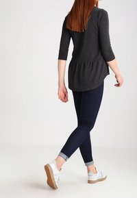 Graues langärmliges Oberteil mit einem Peplum-Detail, kombiniert mit dunkelblauben Skinny Jeans und weißen Sneakers mit silbernen Akzenten, von hinten betrachtet.