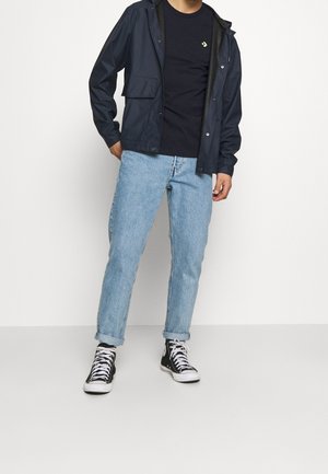 Man draagt een marineblauw jack over een zwart T-shirt, lichtblauwe opgerolde jeans en zwarte hoge sneakers, staand tegen een witte achtergrond.