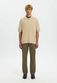 Jeune homme aux cheveux blonds courts, debout face à l'avant, portant un polo beige à manches courtes, un pantalon kaki et des chaussures habillées noires sur un fond blanc.