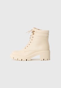 Bottines de couleur crème en cuir lisse avec un poignet côtelé, un talon épais et un laçage à l'avant avec des œillets dorés.