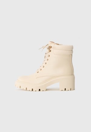 Kremowe ankle boots wykonane z gładkiej skóry, z karbowanym mankietem, masywnym obcasem i sznurowanym przodem z złotymi oczkami.