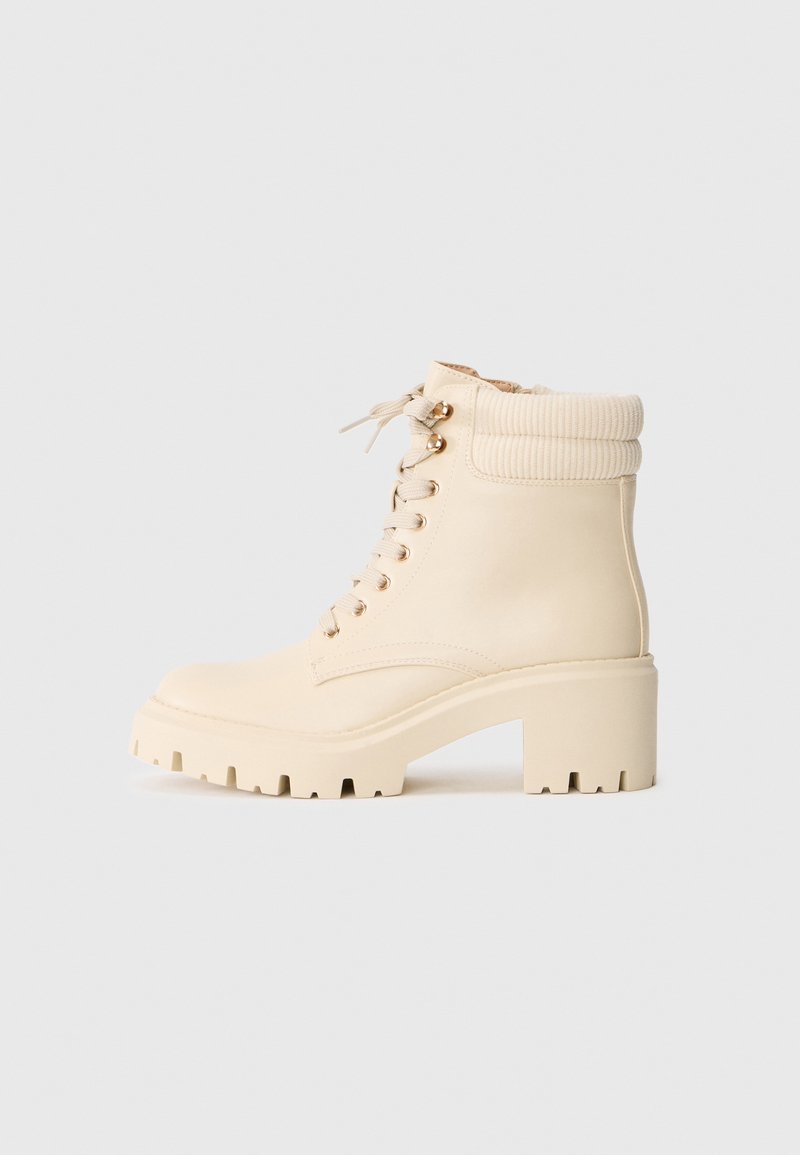 Bottines de couleur crème en cuir lisse avec un poignet côtelé, un talon épais et un laçage à l'avant avec des œillets dorés.
