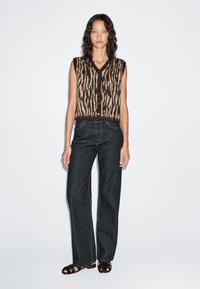 Gebreid mouwloos topje met een bruin en crème dierlijk patroon, knoopsluiting, gecombineerd met hoge donkere spijkerbroek en bruine sandalen.