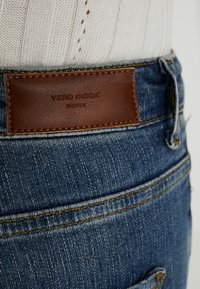 Närbild på blå denimjeans med en brun läderlapp märkt "Vero Moda Denim" ovanför bakfickan och en vit stickad tröja.