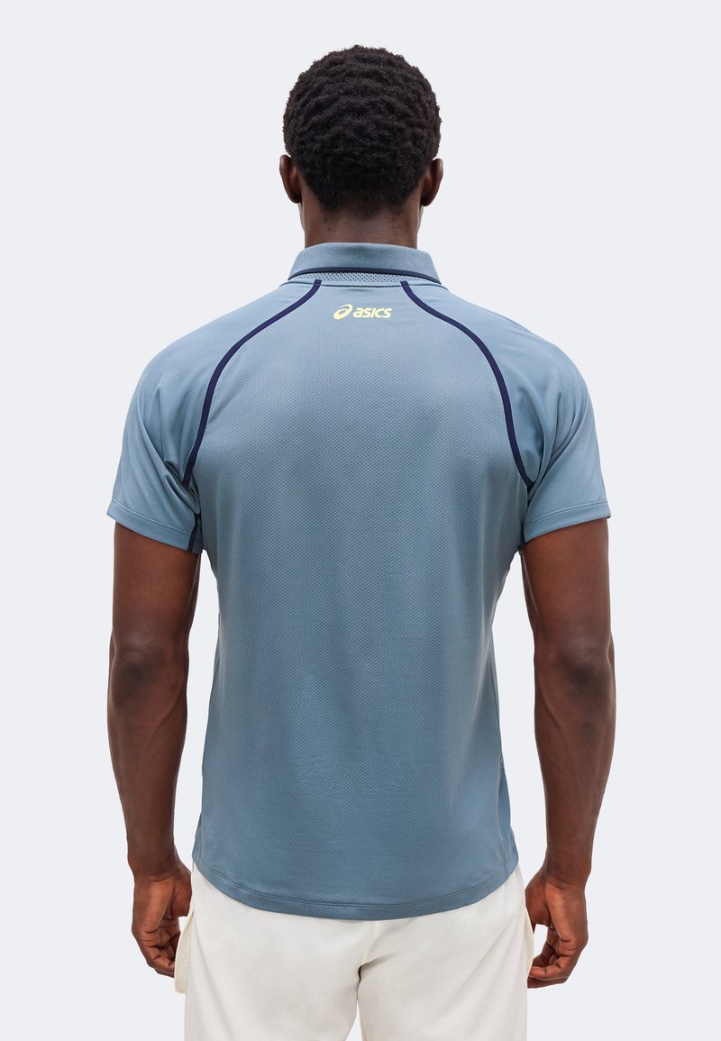 Mann trägt hellblaues kurzärmliges Poloshirt mit dunkelblauen Nähten und gelbem Asics-Logo auf dem oberen Rücken, steht vor einem einfarbigen Hintergrund.