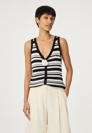 Vrouw draagt zwart-wit gestreepte, mouwloze gehaakte top met diepe V-hals en crème wijde broek, staand tegen een effen achtergrond.