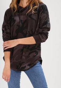 Sudadera con patrón de camuflaje oscuro hecha de una tela suave, con capucha ajustable y un corte relajado, combinada con unos jeans de color azul claro.