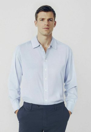 Chemise à manches longues bleu clair, à col, en tissu texturé, avec des boutons blancs, associée à un pantalon sombre. Design simple.