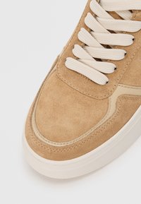 Baskets en suede beige avec un bout arrondi, des accents beiges et des lacets crème. Texture lisse avec des détails de couture visibles.