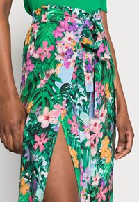 Jupe enveloppante florale avec des motifs de fleurs multicolores sur fond vert vif ; fabriquée en tissu léger, dotée d'une fente sur le côté et d'une taille à nœud.