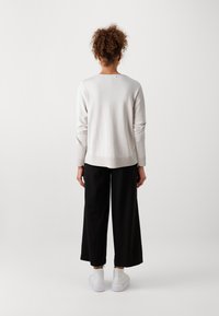 Lys grå langermet genser med avslappet passform, parret med svarte vide bukser og hvite sneakers, sett fra bak.