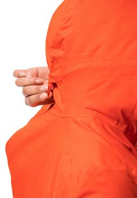 Oranžová nepromokavá bunda s stahovací kapucí. Hladká textura, viditelné švy a ruka upravující šňůrku na kapuci.