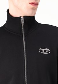 Casaco preto com fecho zip, com um colarinho alto, feito de um material suave. Apresenta um logótipo prateado no lado esquerdo do peito e um fecho metálico.