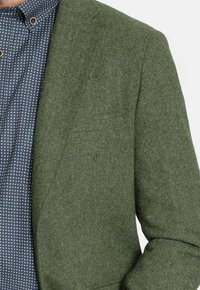 Blazer verde texturizado con solapa de muesca, combinado con una camisa azul con lunares. Presenta un bolsillo en el pecho y detalles de botones en el cuello.
