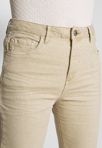 Jeans beige de corte slim, con frente plano, cinco bolsillos y cierre de botón en la cintura. Textura suave con detalles de costura sutiles.