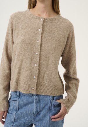 Cárdigan beige de punto con botones de perla, usado con vaqueros azules, mano descansando en el bolsillo, fondo neutro.
