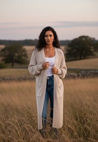 Beżowy sweter kardigan o długim kroju, z prążkowanymi rękawami i dwoma przednimi kieszeniami, noszony na białej bluzce i niebieskich dżinsach.