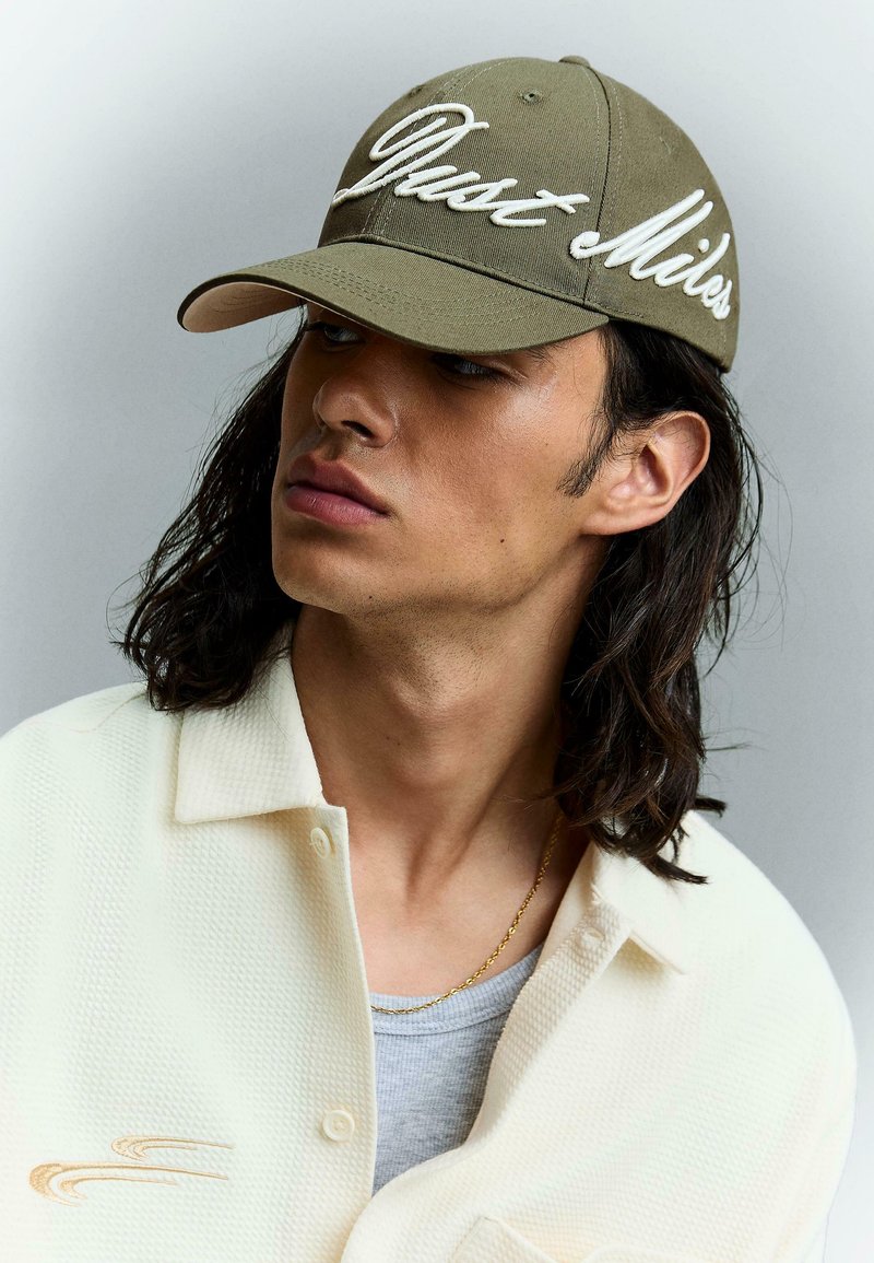 Jeune homme aux cheveux noirs mi-longs portant une casquette vert olive avec une broderie blanche et une chemise crème boutonnée par-dessus un haut gris.