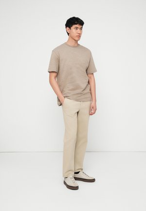 Young man standing with hands in pockets wearing beige striped t-shirt, light beige pants, and beige sneakers with brown soles.