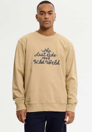 Joven con sudadera beige con texto oscuro "The Last Ride 1949 Wild World", de pie frente a un fondo blanco liso.