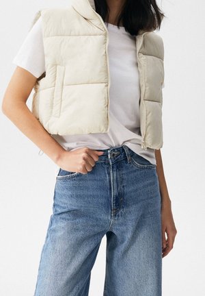 Persoon draagt een lichtbeige gewatteerd vest over een wit t-shirt en hoog gevormde blauwe spijkerbroek, staand met één hand bij de taille.