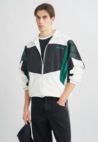 V白s un melnas krūzītes jakas ar zaļām svītrām, elastīgām aprocēm un logotipu uz krūšu daļas, apvienotas ar melnām džinsām.