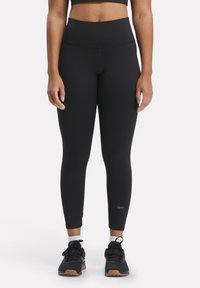 Leggings neri a vita alta con una texture liscia, un design aderente e un dettaglio riflettente del logo vicino alla caviglia. Indossati con scarpe da ginnastica nere.