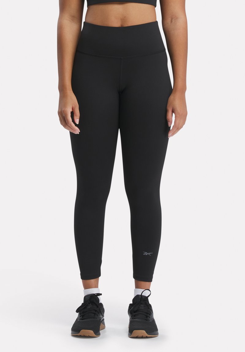 Leggings neri a vita alta con una texture liscia, un design aderente e un dettaglio riflettente del logo vicino alla caviglia. Indossati con scarpe da ginnastica nere.