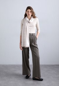 Haut beige à manches longues, pantalons gris à larges jambes et une écharpe à rayures claires avec des franges. Des chaussures plates noires complètent la tenue. Fond uni.