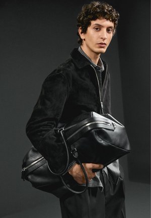Mladý muž v černé bundě drží velkou černou koženou sportovní tašku na tmavém pozadí.
