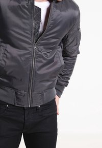 Veste bomber grise en nylon lisse avec fermeture éclair, poignets et taille côtelés. Comprend une poche latérale sur la manche.