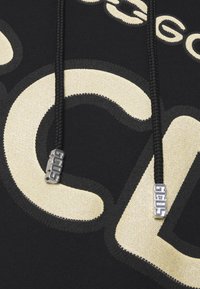 Felpa nera con grandi lettere color crema e accenti testurizzati. Presenta lacci neri con punte metalliche che mostrano il nome del marchio.