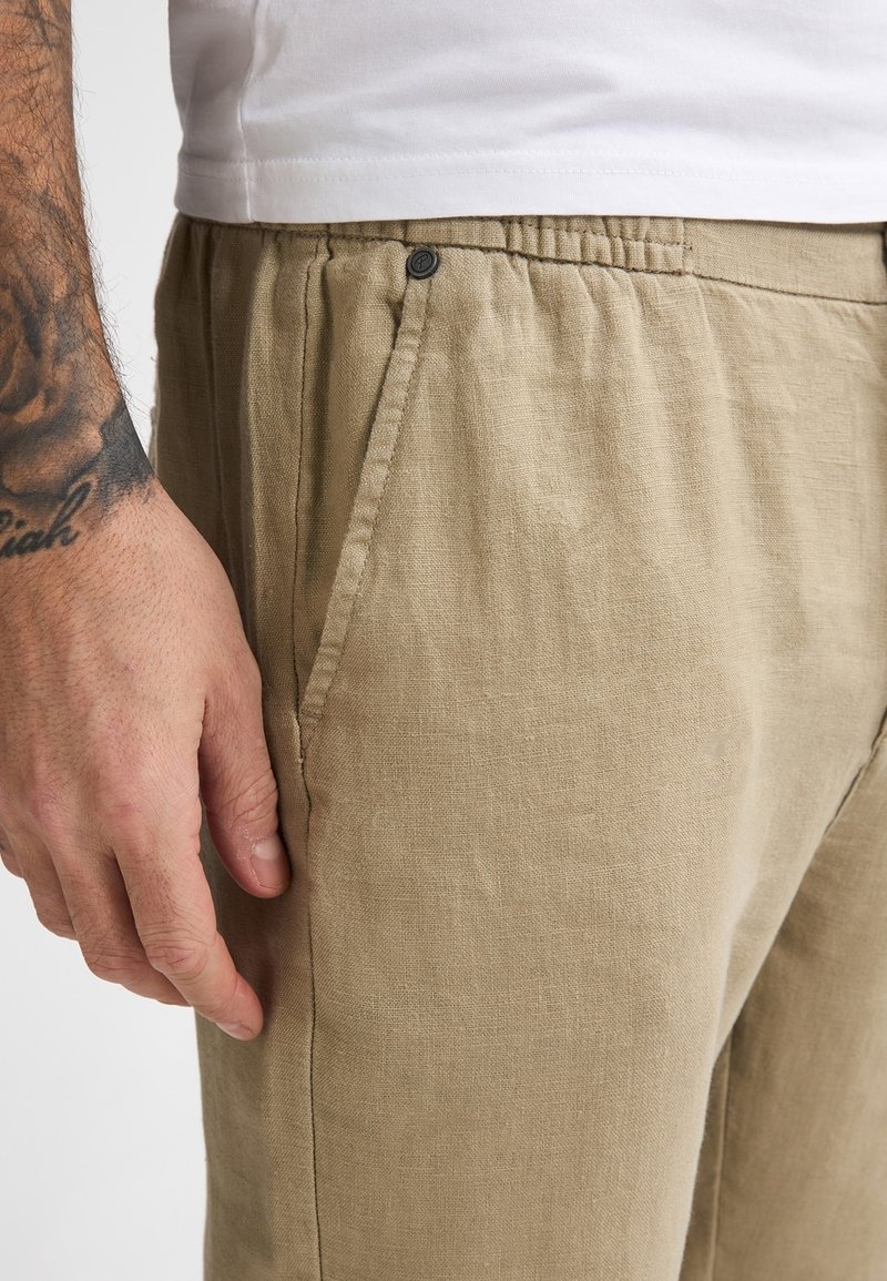 Pantalon en lin beige avec taille élastique, poches latérales et détail en bouton près de la taille. Tissu texturé visible.