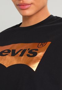 Musta t-paita pyöreällä kaula-aukoilla, jossa on suuri metallinhohtoinen kuparinvärinen graafinen logo. Pehmeää puuvillamateriaalia, jossa on sileä pinta.