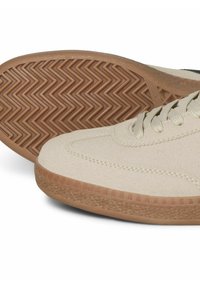 Baskets en suède beige avec une semelle en caoutchouc texturé présentant un motif en chevrons, des lacets plats et des accents noirs contrastants sur le dessus.