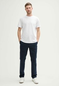 Camiseta de algodón blanca de manga corta, combinada con chinos azul marino y zapatillas blancas, de pie sobre un fondo claro y liso.