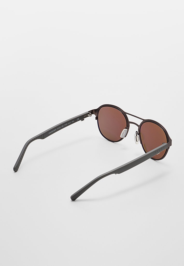 Gafas de sol de montura redonda en negro mate con lentes marrones y almohadillas nasales ajustables, descansando sobre una superficie blanca.