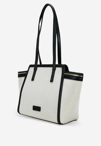 Bolsa tote con un cuerpo de lienzo texturizado en crema, detalles en cuero negro y dos asas para el hombro; cuenta con un cierre con cremallera y un detalle del logo.