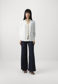 Blusa bianca a maniche lunghe con dettagli in zip dorata e polsini larghi, abbinata a jeans a zampa d'elefante blu scuro e sandali neri con tacco alto.