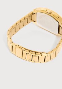 Bracciale per orologio in metallo dorato con fibbia, collegato a una cassa quadrata dell'orologio, mostrato dal retro su uno sfondo bianco.