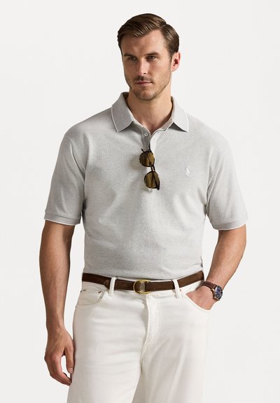 Graues Polohemd mit kurzen Ärmeln und Kragen, mit einem kleinen Logo auf der Brust. Getragen mit weißen Hosen und einem braunen Gürtel. Eine Brille liegt auf dem Hemd.
