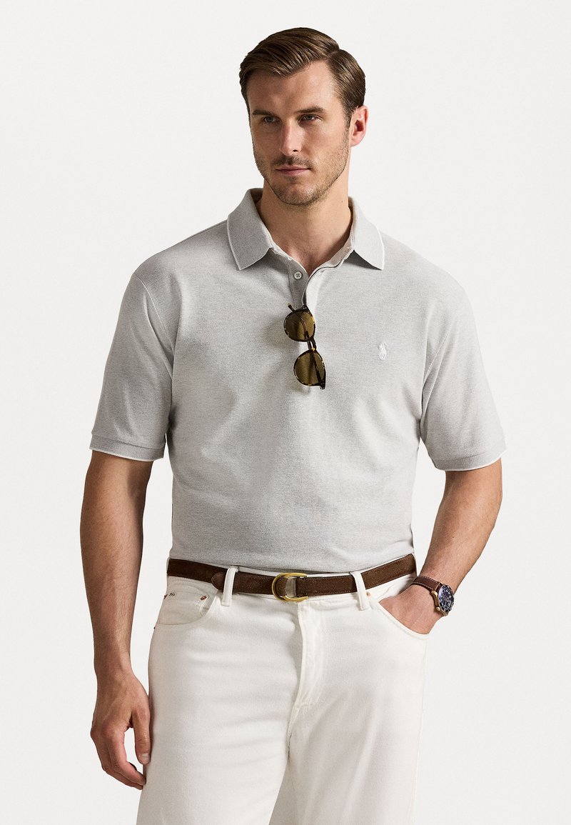 Siva kratka polo majica s ovratnikom, ki ima majhen logotip na prsih. Obuta z belimi hlačami in rjavim pasom. Očala ležijo na majici.