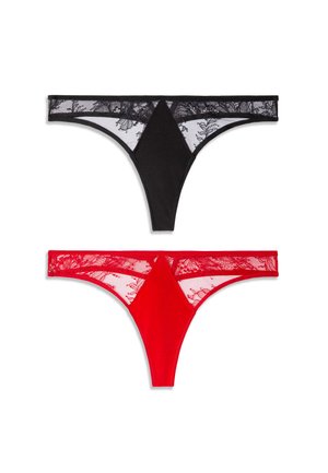 Femme aux longs cheveux foncés porte un soutien-gorge en dentelle noire transparente et une culotte assortie, debout pieds nus devant un fond clair uni.