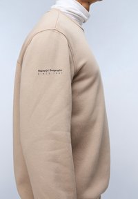 Sudadera beige con cuello y puños acanalados. Textura suave. Presenta texto de logo en la manga izquierda con "Napapijri Geographic SINCE 1987."