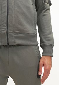 Felpa grigia con zip, dotata di polsini e orlo a costine, con una texture liscia. Presenta cuciture visibili e tasche di accesso sui pantaloni coordinati.
