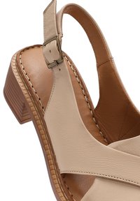 Sandali con tacco in pelle beige con design aperto, cinturino alla caviglia regolabile e dettagli cuciti su una suola marrone texturizzata.