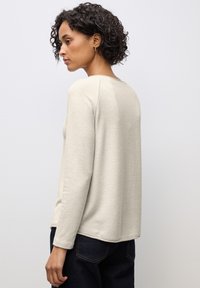 Helles beige Langarmshirt aus einem weichen, strukturierten Stoff mit lässiger Passform und rundem Ausschnitt, kombiniert mit dunkelblauen Jeans.