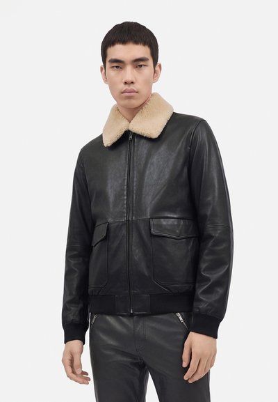 Jeune homme portant une veste en cuir noire avec un col en shearling beige et un pantalon en cuir noir assorti sur un fond uni.