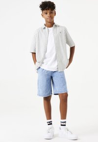 Gestreepte kortormige overhemden in wit en grijs, gecombineerd met spijkerbroekshorts en witte sneakers. Zwarte en witte gestreepte sokken zichtbaar.