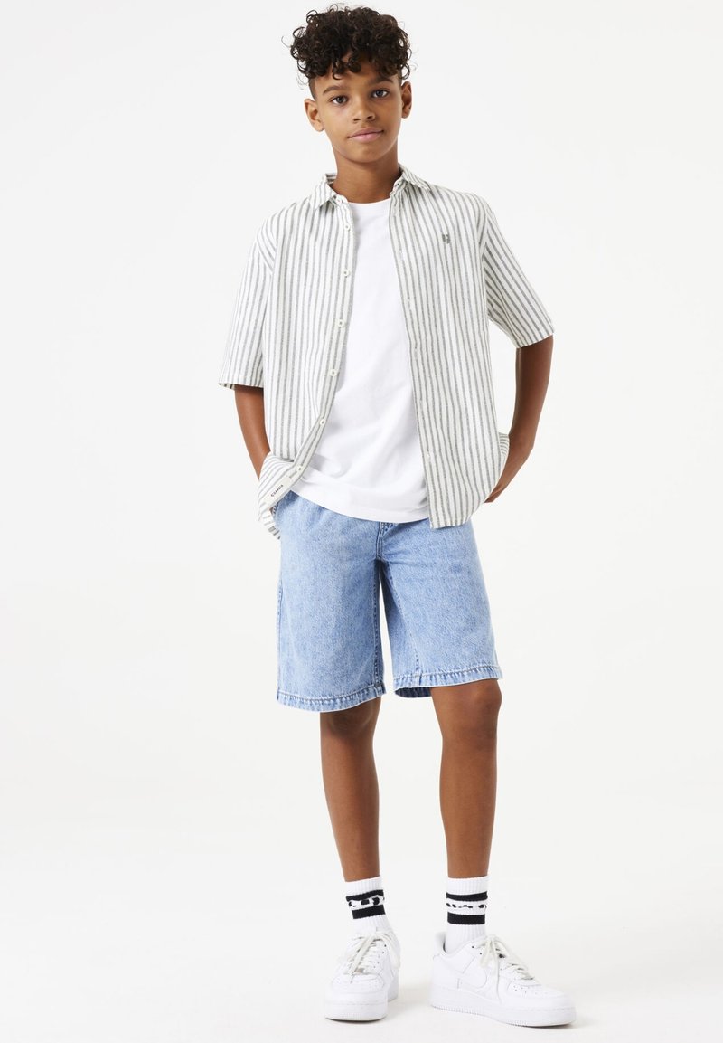 Gestreepte kortormige overhemden in wit en grijs, gecombineerd met spijkerbroekshorts en witte sneakers. Zwarte en witte gestreepte sokken zichtbaar.